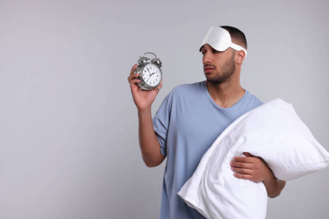 Man in sleepwear holding clock and pillow with tired look