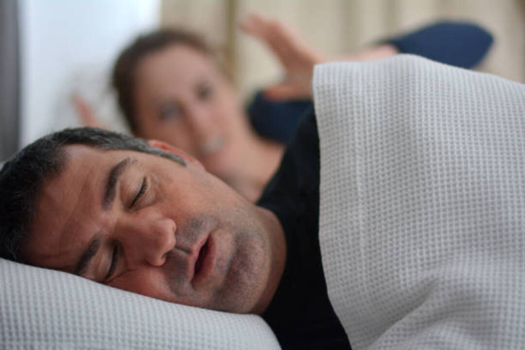 Man snoring loudly in bed while woman sleeps in background.