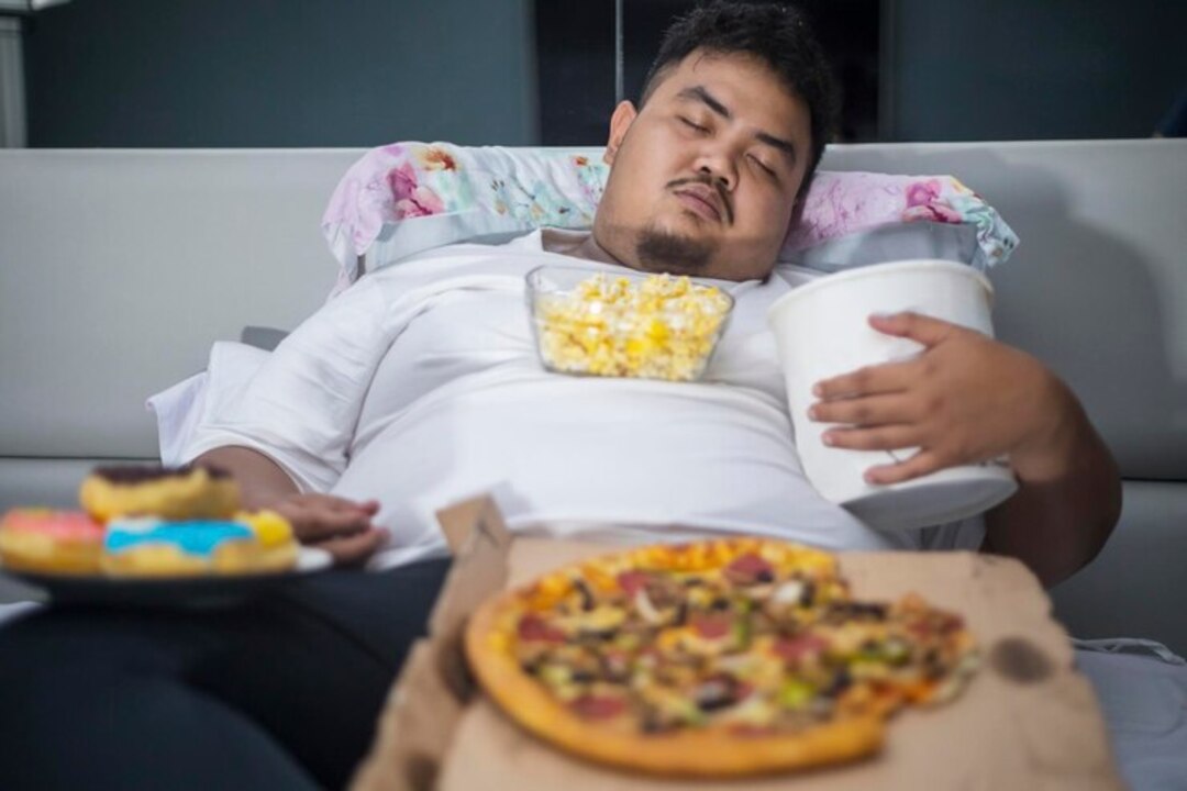 Overweight man asleep surrounded by junk food and snacks.