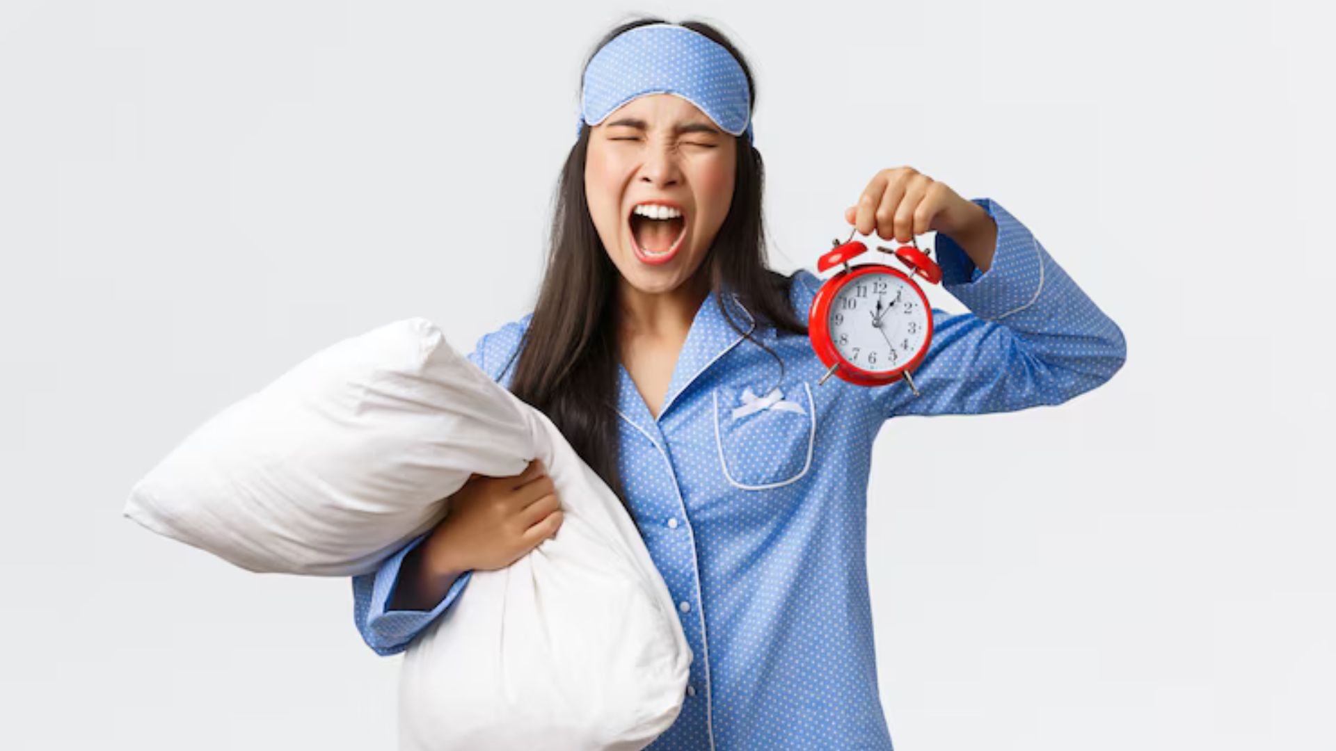 Woman in pajamas holding a pillow and alarm clock, frustrated.