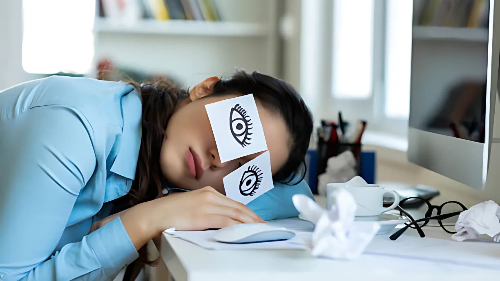 Woman resting at a desk with paper eyes, showing sleepiness.